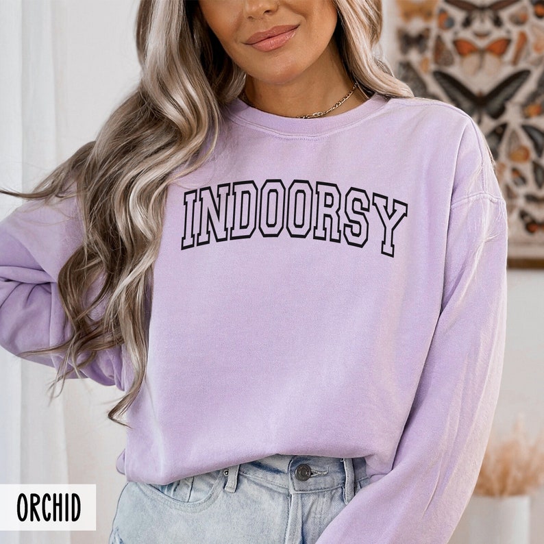 Pode incluir: Uma camisola de manga comprida de cor lavanda com o texto "INDOORSY" impresso a preto na frente. A camisola &eacute; feita de um material macio e confort&aacute;vel.