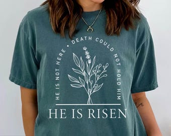 Puede incluir: Una camiseta azul turquesa con un diseño gráfico blanco. El gráfico presenta un diseño floral con el texto "Death could not hold him. He is not here. He is risen."