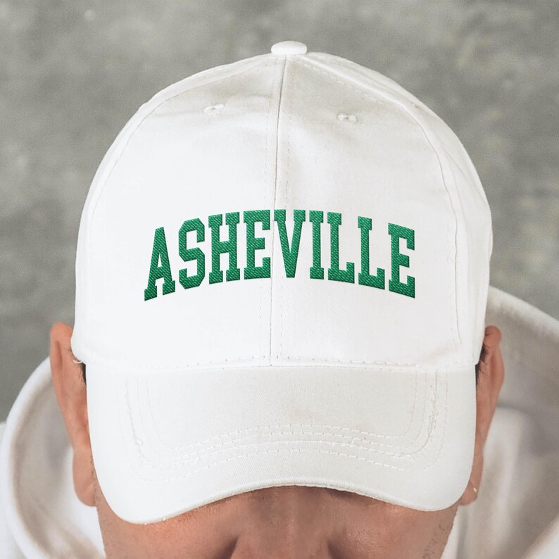 Puede incluir: Gorra de b&eacute;isbol blanca con la palabra "ASHEVILLE" bordada en letras verdes en la parte delantera. La gorra tiene una visera curva y un bot&oacute;n en la parte superior. El fondo es gris neutro.
