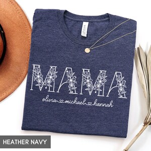 May include: Heather navy t-shirt with the word "MAMA" in white floral letters. Below the word "MAMA" are the names "olivia michael hannah" in white script. A gold necklace with a round pendant is on the shirt.