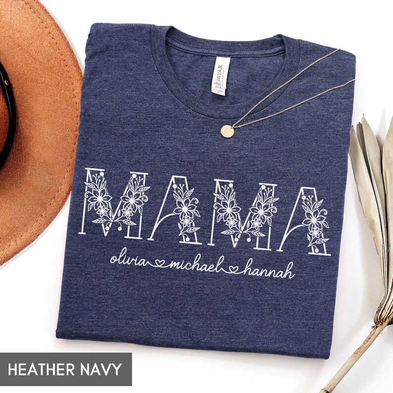 May include: Heather navy t-shirt with the word "MAMA" in white floral letters. Below the word "MAMA" are the names "olivia michael hannah" in white script. A gold necklace with a round pendant is on the shirt.