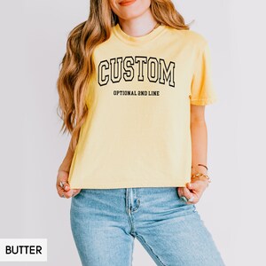 May include: A yellow cropped t-shirt with the word "CUSTOM" in black outlined letters. The text "OPTIONAL 2ND LINE" is printed below the word "CUSTOM".