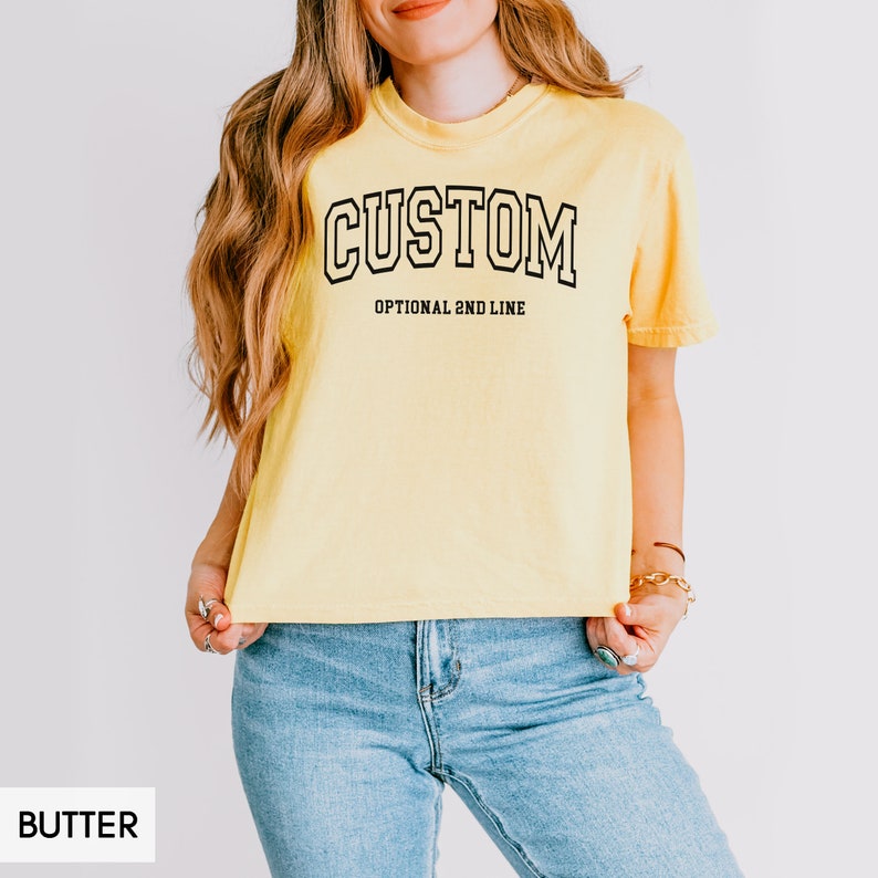 May include: A yellow cropped t-shirt with the word "CUSTOM" in black outlined letters. The text "OPTIONAL 2ND LINE" is printed below the word "CUSTOM".