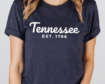 May include: A heather navy short-sleeve t-shirt with rolled sleeves. The front features white text that reads Tennessee in a cursive script font, with the text EST. 1796 printed in a clean, sans-serif font directly underneath the state name.