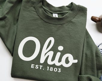 May include: Military green sweatshirt with the word "Ohio" in white script, and "EST. 1803" below. The sweatshirt is a solid color and has a crew neck. The bottom of the image shows a pair of blue jeans.
