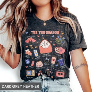 May include: A dark grey heather t-shirt featuring the text 'TIS THE SEASON and various Valentine's Day themed illustrations including an open envelope, heart balloons, flowers, a box of chocolates, a calendar, candles, jewelry, and a cassette tape.