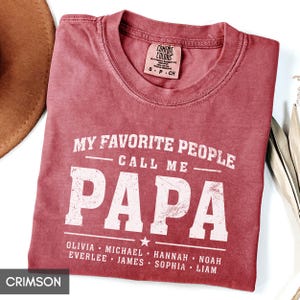 A red t-shirt with the text "MY FAVORITE PEOPLE CALL ME PAPA" printed on it, along with the names "OLIVIA, MICHAEL, HANNAH, NOAH, EVERLEE, JAMES, SOPHIA, Liam" printed below.