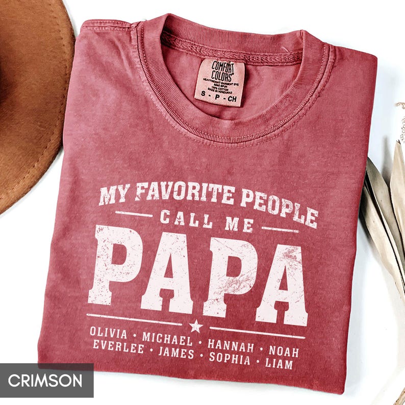 A red t-shirt with the text "MY FAVORITE PEOPLE CALL ME PAPA" printed on it, along with the names "OLIVIA, MICHAEL, HANNAH, NOAH, EVERLEE, JAMES, SOPHIA, Liam" printed below.