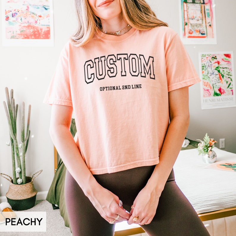 May include: Peachy colored cropped t-shirt with the word "CUSTOM" in a black outlined, white filled, varsity style font. The text "OPTIONAL 2ND LINE" is printed below the word "CUSTOM".