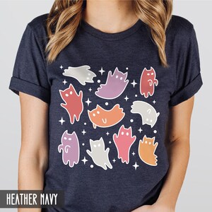 Op de afbeelding: Een heideblauw t-shirt met een kleurrijk spookkattenontwerp. De katten zijn in verschillende tinten roze, paars, oranje en grijs. Het ontwerp is omringd door witte sterren.