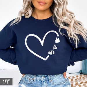 May include: Navy blue sweatshirt with a white outline of a heart, a camper, a campfire, and trees inside the heart. The text "NAVY" is in the bottom left corner of the image.