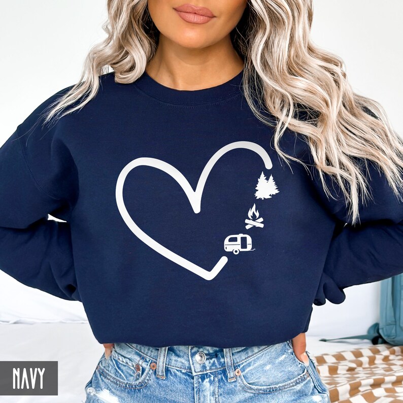 May include: Navy blue sweatshirt with a white outline of a heart, a camper, a campfire, and trees inside the heart. The text "NAVY" is in the bottom left corner of the image.