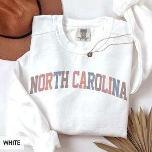 Puede incluir: Sudadera blanca de cuello redondo con el texto "NORTH CAROLINA" en un dise&ntilde;o arqueado multicolor. La sudadera tiene una etiqueta que dice "COMFORT COLORS". Un collar dorado con un colgante est&aacute; drapeado sobre la sudadera.