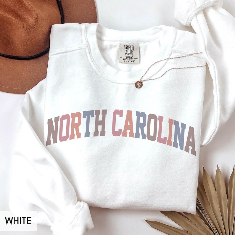 Puede incluir: Sudadera blanca de cuello redondo con el texto "NORTH CAROLINA" en un dise&ntilde;o arqueado multicolor. La sudadera tiene una etiqueta que dice "COMFORT COLORS". Un collar dorado con un colgante est&aacute; drapeado sobre la sudadera.