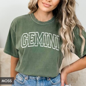 May include: A moss green t-shirt with the word "GEMINI" printed in white block letters. The shirt has a crew neck and short sleeves. The word "MOSS" is in a small box in the bottom left corner.