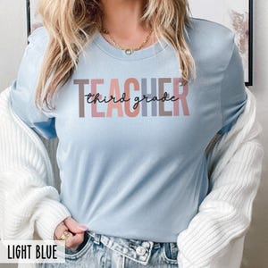 A woman wearing a light blue t-shirt with multicolored block letters spelling teacher and black cursive text reading third grade.