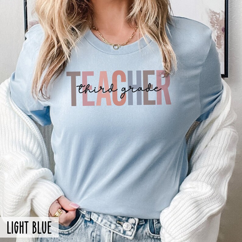 A woman wearing a light blue t-shirt with multicolored block letters spelling teacher and black cursive text reading third grade.