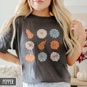 May include: A dark gray t-shirt with a graphic print of nine different pumpkins in various colors and styles. The pumpkins are arranged in a grid pattern. The word "PEPPER" is printed in white at the bottom of the shirt.