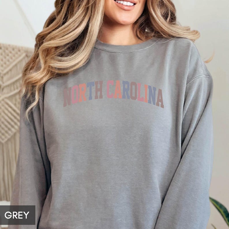 Puede incluir: Una sudadera gris con el texto "NORTH CAROLINA" en un dise&ntilde;o arqueado multicolor. La sudadera es de color gris liso y tiene mangas largas. La palabra "GREY" est&aacute; en un peque&ntilde;o cuadro en la esquina inferior izquierda.