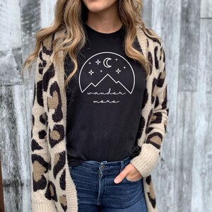 A woman wearing a black wander more t-shirt under a leopard-print cardigan and blue jeans poses in front of a gray wooden wall.