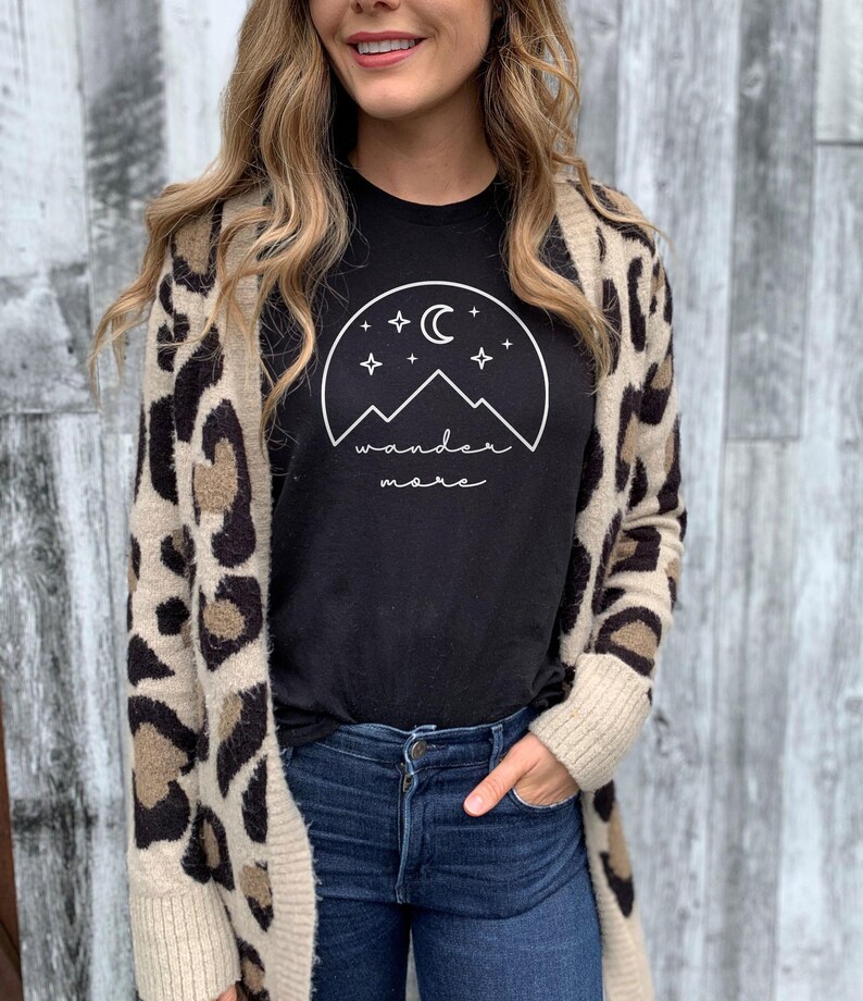 A woman wearing a black wander more t-shirt under a leopard-print cardigan and blue jeans poses in front of a gray wooden wall.