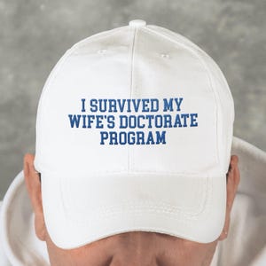May include: White baseball cap with the text "I SURVIVED MY WIFE'S DOCTORATE PROGRAM" in blue lettering. The cap has a curved brim and a button on top. The hat is made of a soft material.