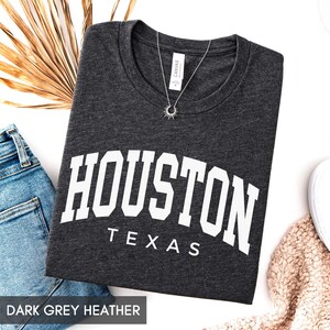 May include: A dark grey heather t-shirt featuring the white text HOUSTON TEXAS in a bold, arched collegiate font. The shirt has a crew neckline and a small white tag inside the collar. A silver sunburst pendant necklace rests on the fabric.