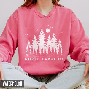 May include: A pink sweatshirt with a white graphic of a forest silhouette and the text "NORTH CAROLINA". The sweatshirt is a crew neck style and has a distressed look.