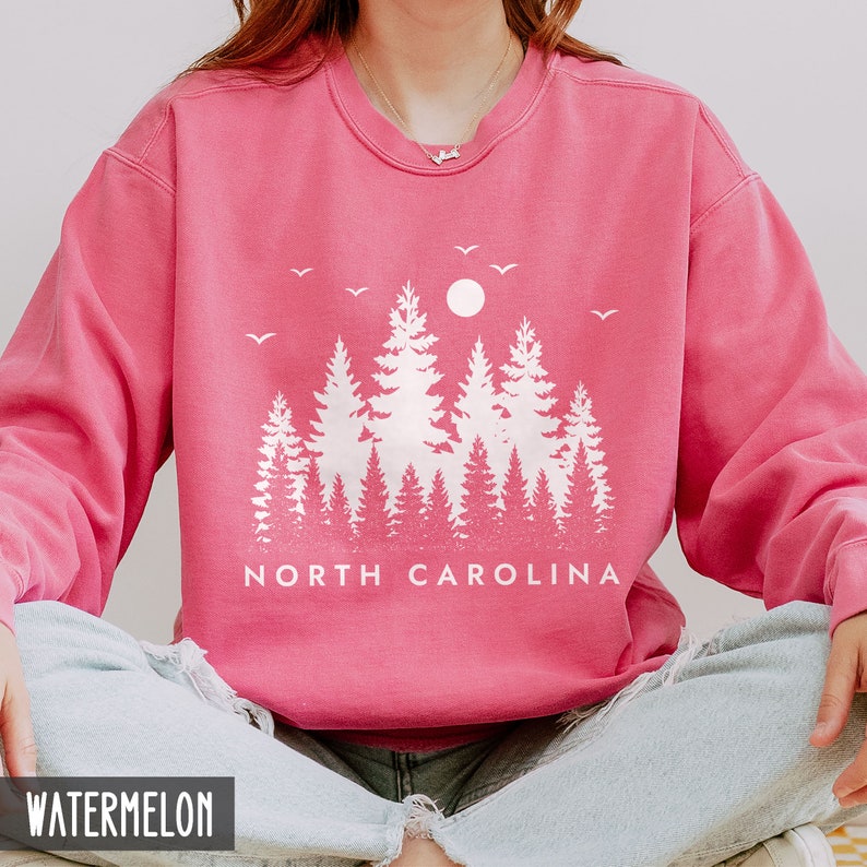 May include: A pink sweatshirt with a white graphic of a forest silhouette and the text "NORTH CAROLINA". The sweatshirt is a crew neck style and has a distressed look.