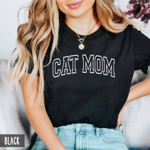 May include: Black t-shirt with white text that reads "CAT MOM" in a varsity style font.