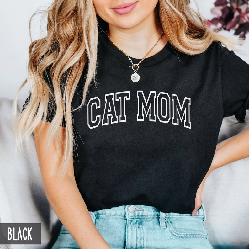 May include: Black t-shirt with white text that reads "CAT MOM" in a varsity style font.