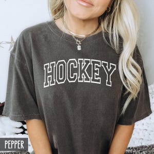 A woman wearing a dark pepper gray t-shirt with white block lettering spelling HOCKEY.