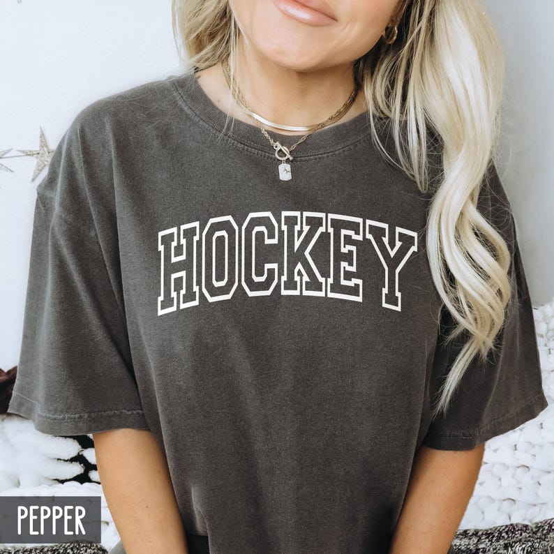 A woman wearing a dark pepper gray t-shirt with white block lettering spelling HOCKEY.