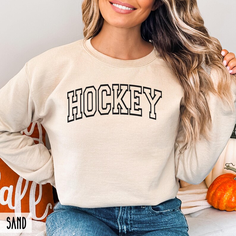 A woman wearing a sand-colored crewneck sweatshirt with the word HOCKEY in black collegiate block letters.