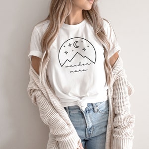 Pode incluir: Camiseta branca com um gr&aacute;fico preto de montanhas, uma lua crescente e estrelas. O texto "wander more" est&aacute; abaixo do gr&aacute;fico.
