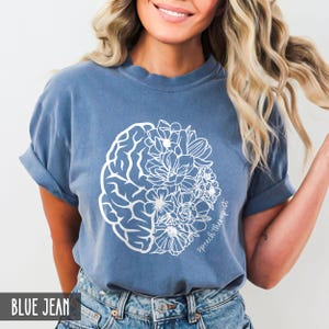 May include: A blue jean t-shirt with a white outline of a brain and flowers. The text "Speech Therapist" is written on the shirt.