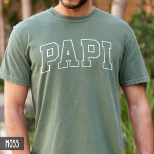 May include: Moss green t-shirt with the word "PAPI" in white, outlined block letters. The shirt has short sleeves and a crew neck. The word "MOSS" is in a small box in the lower left corner.