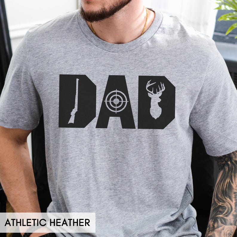 May include: Athletic heather gray t-shirt with the word "DAD" in bold black letters. The "D" features a rifle, the "A" a target, and the second "D" a deer head. The shirt is made of a soft, comfortable material.