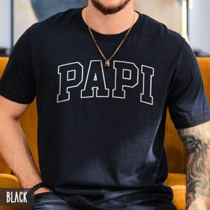 May include: A black short-sleeved t-shirt featuring the word PAPI printed in a large white collegiate-style font across the chest. The shirt has a crew neckline and is made of a solid black fabric. The text is centered and clearly visible.