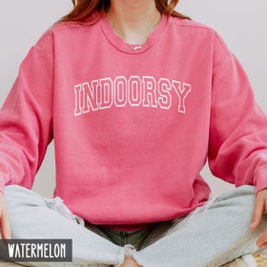 Pode incluir: Uma camisola rosa com o texto "INDOORSY" em letras brancas com contorno. A camisola &eacute; feita de um material macio e confort&aacute;vel e tem um corte descontra&iacute;do.