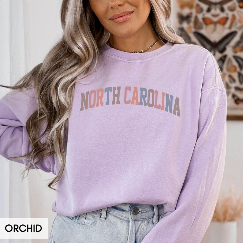 Puede incluir: Sudadera color orqu&iacute;dea con el texto "NORTH CAROLINA" en una fuente retro multicolor. La parte superior de manga larga es de color liso y tiene cuello redondo. La palabra "ORCHID" est&aacute; en un cuadro blanco en la parte inferior izquierda.
