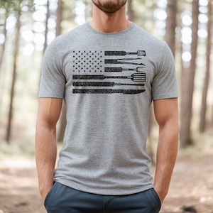 Puede incluir: Una camiseta gris con un dise&ntilde;o de bandera estadounidense en blanco y negro. La bandera est&aacute; formada por utensilios de cocina, como una esp&aacute;tula, pinzas y un cuchillo.