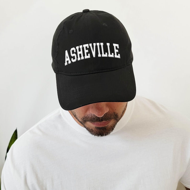 Puede incluir: Gorra de b&eacute;isbol negra con la palabra "ASHEVILLE" bordada en blanco en la parte delantera. La gorra la lleva una persona que lleva una camiseta blanca. El fondo es blanco liso.