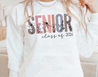 May include: White sweatshirt with the word "SENIOR" in a multi-colored block letter design, with floral accents. Below the word "SENIOR" is the phrase "class of 2026" in a handwritten font. The sweatshirt is a crew neck style.
