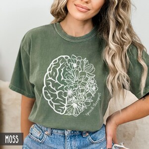 May include: A moss green t-shirt with a white outline of a brain and flowers. The text "Speech Therapist" is written on the shirt.