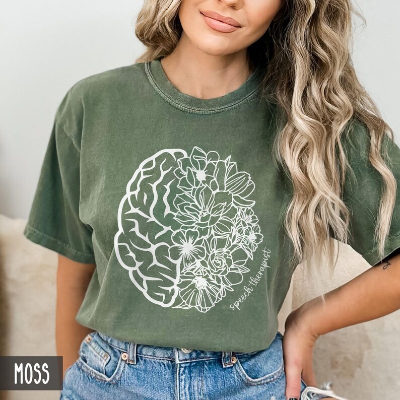 May include: A moss green t-shirt with a white outline of a brain and flowers. The text "Speech Therapist" is written on the shirt.
