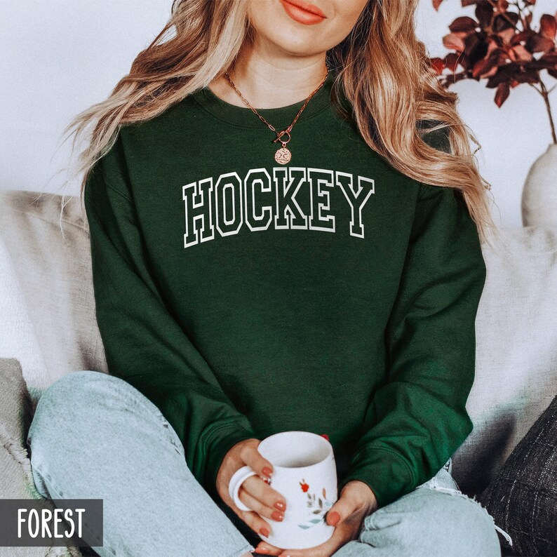 A woman sitting on a couch holding a mug, wearing a forest green crewneck sweatshirt with the word HOCKEY in white collegiate block letters.