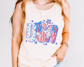 May include: Ivory-colored tank top with a patriotic "USA" graphic. The graphic features red, white, and blue stars and stripes, with a blue bow. The tank top is sleeveless and made of a soft material.