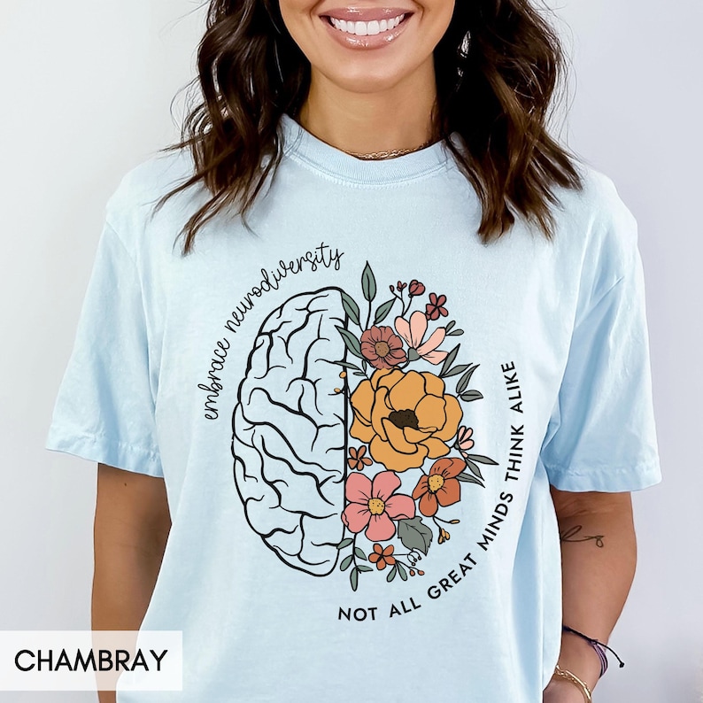以下が含まれることがあります： 薄い青色のTシャツに、花が咲いた脳のグラフィックと「Embrace neurodiversity. Not all great minds think alike.」というテキストがプリントされています