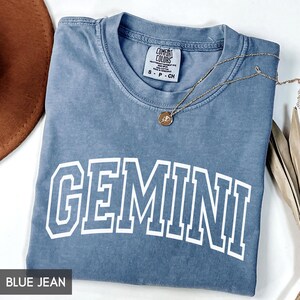 May include: Blue jean colored t-shirt with the word "GEMINI" in white block letters. The shirt has a crew neck and a small tag at the neckline. A gold necklace with a pendant is draped across the shirt.